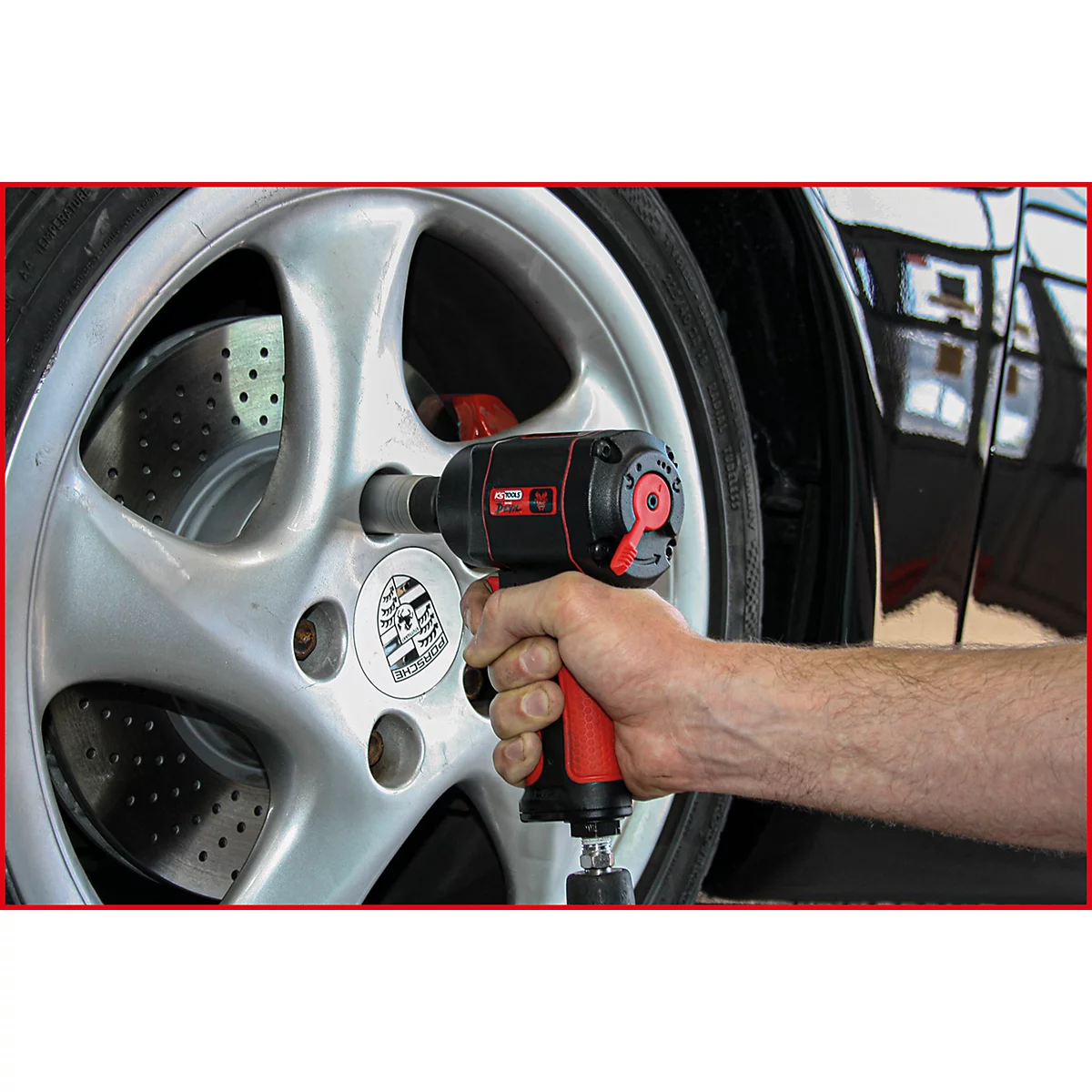 Ein Mann hält einen schwarzen und roten Schlagschrauber vor einem silbernen Porsche-Rad. Das Rad weist ein Porsche-Emblem auf.